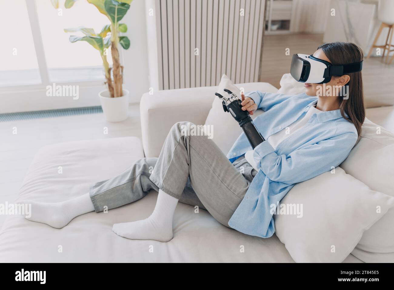 Relaxed young woman with a prosthetic arm enjoys a virtual reality experience, comfortably lounging at home Stock Photo