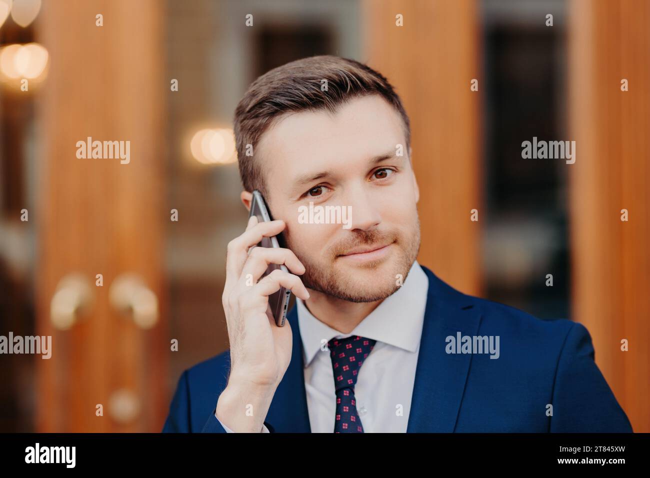 Professional man in blue suit on phone, confident gaze, wooden doors Stock Photo