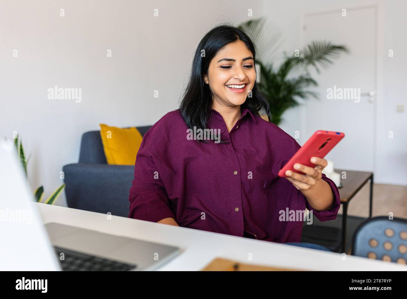 Happy indian woman using mobile phone sitting with laptop on table at home Stock Photo