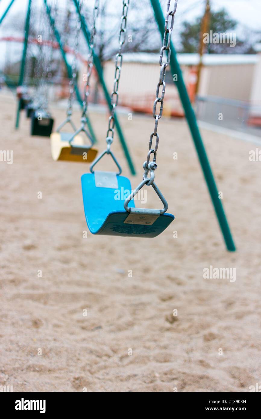 A row of multicolored swings on a playground, with a large sand-covered play area, and portable buildings in the background. Stock Photo