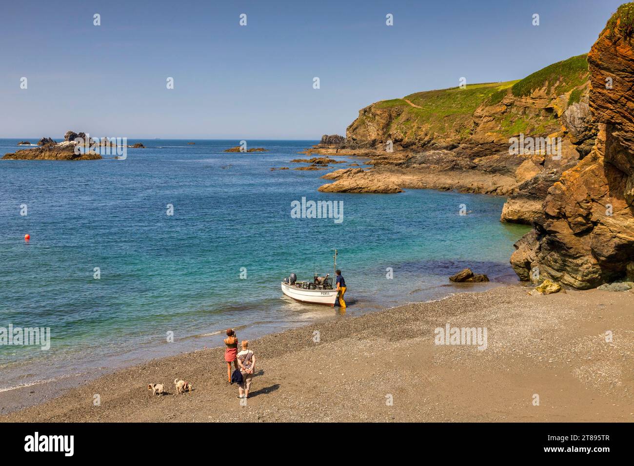 22 May 2023: Lizard Cove, Cornwall, UK - Small cove at The Lizard, with man launching a boat and tourist with two dogs. Perfect spring day. Stock Photo