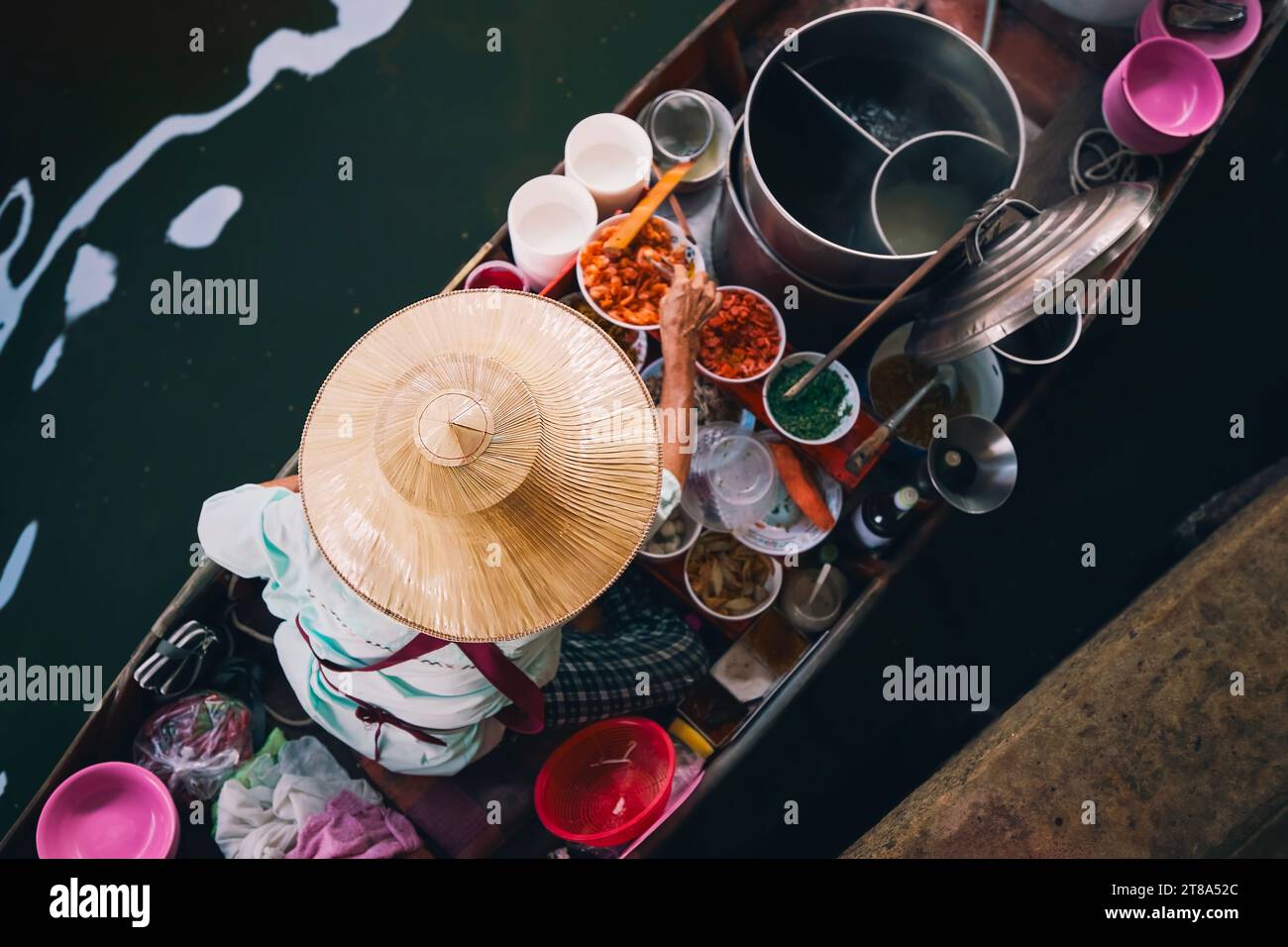 Selective focus on traditional hat of vendor on boat on floating market. Woman is preparing food on water in Damnoen Saduak near Bangkok, Thailand. Stock Photo