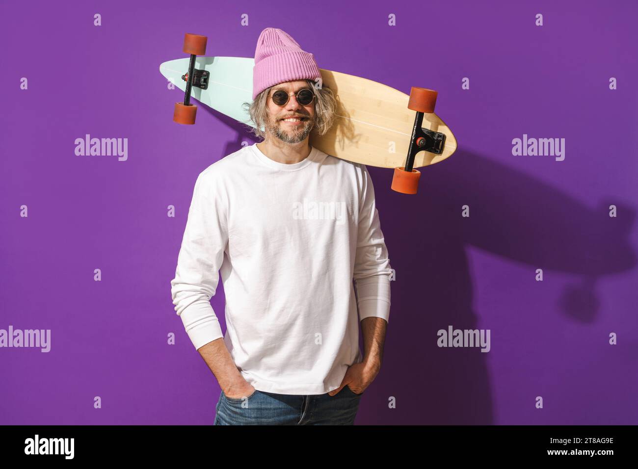 Cheerful middle aged man with longboard wearing white sweatshirt against purple background Stock Photo