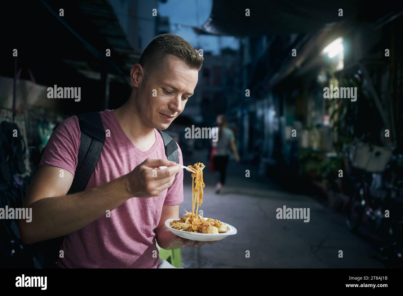 Tourist at night Bangkok. Man with backpack eating local food (pad thai) at street market. Stock Photo