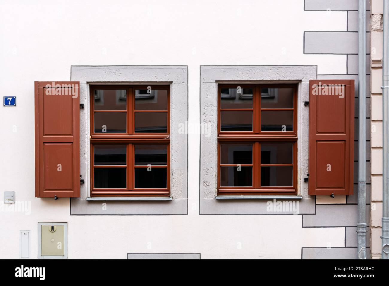 Two rectangular windows with brown frames and wooden shutters against a white wall with a drainpipe. From the Windows of the world series. Stock Photo