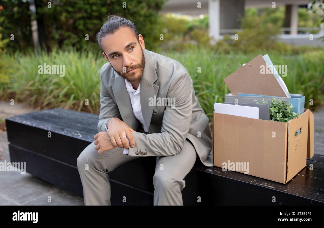 Upset young office employee sitting with box of personal stuff outdoors Stock Photo