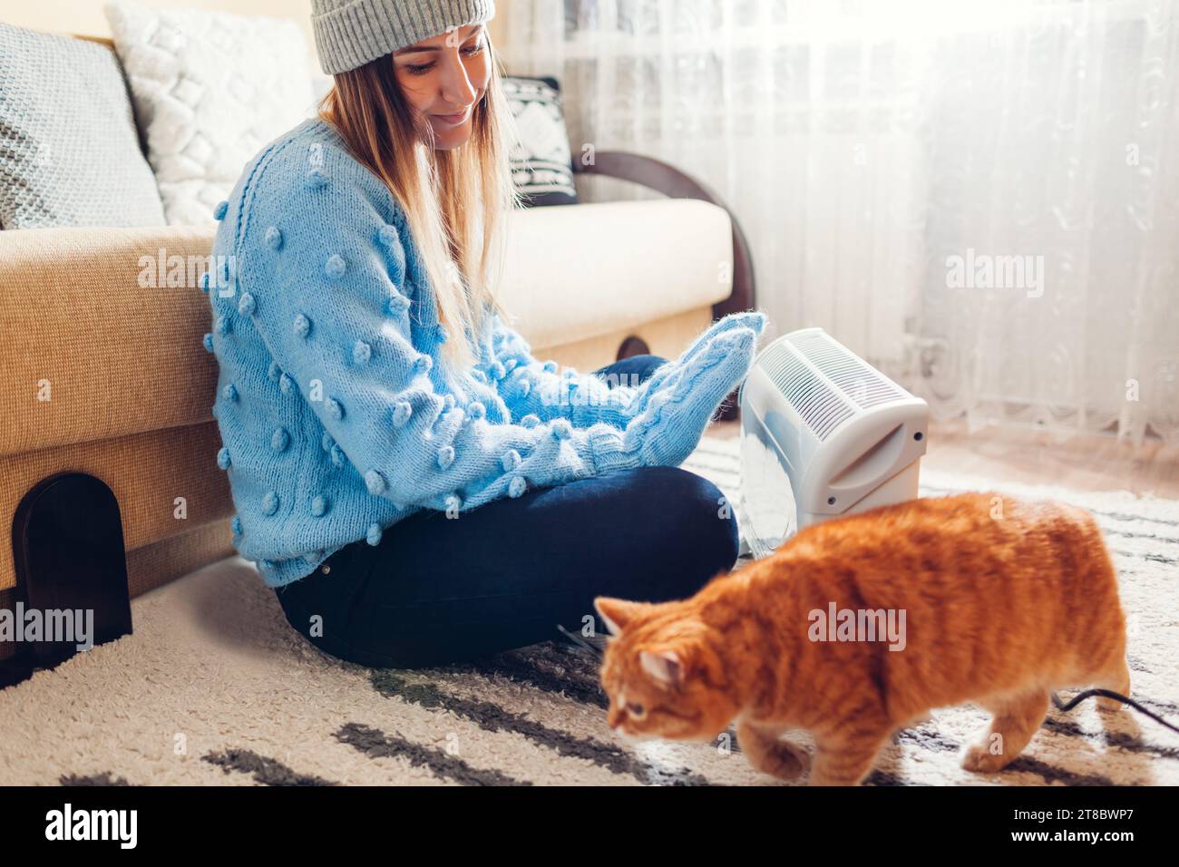 Using heater at home in winter. Woman warming her hands with cat wearing sweater. Heating season. Stock Photo