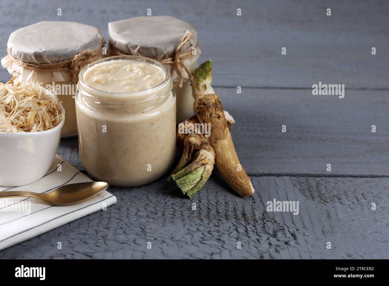 Spicy horseradish sauce in jars, roots and spoon on grey wooden table