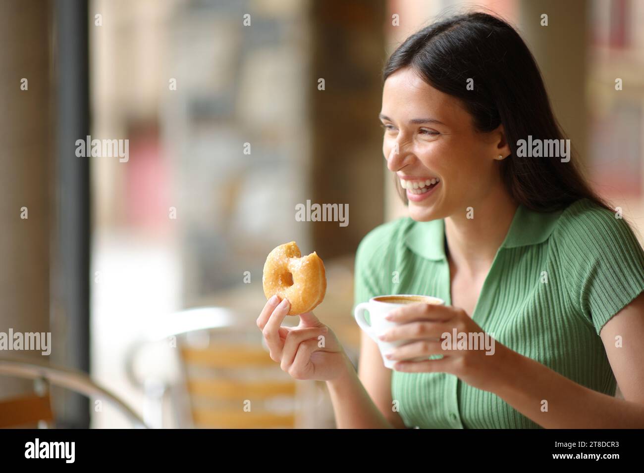 Happy woman laughing at breakfast in a bar terrace eating doughnut and drinking coffee Stock Photo