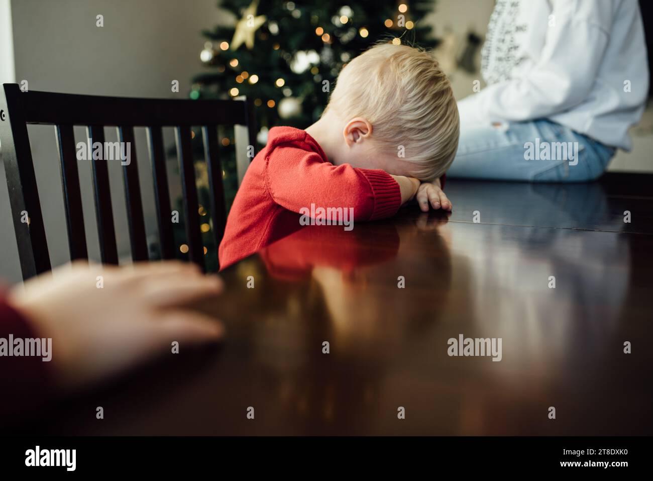 Little boy sits with head down on table pouting in front of Chri Stock Photo