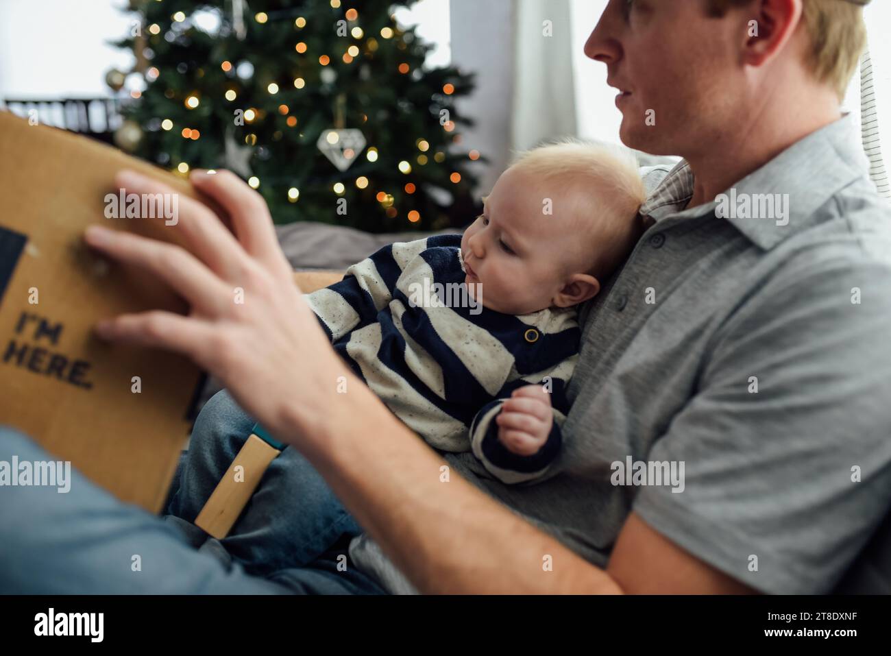 Baby boy sits on dad's lap while opening christmas presents in f Stock Photo