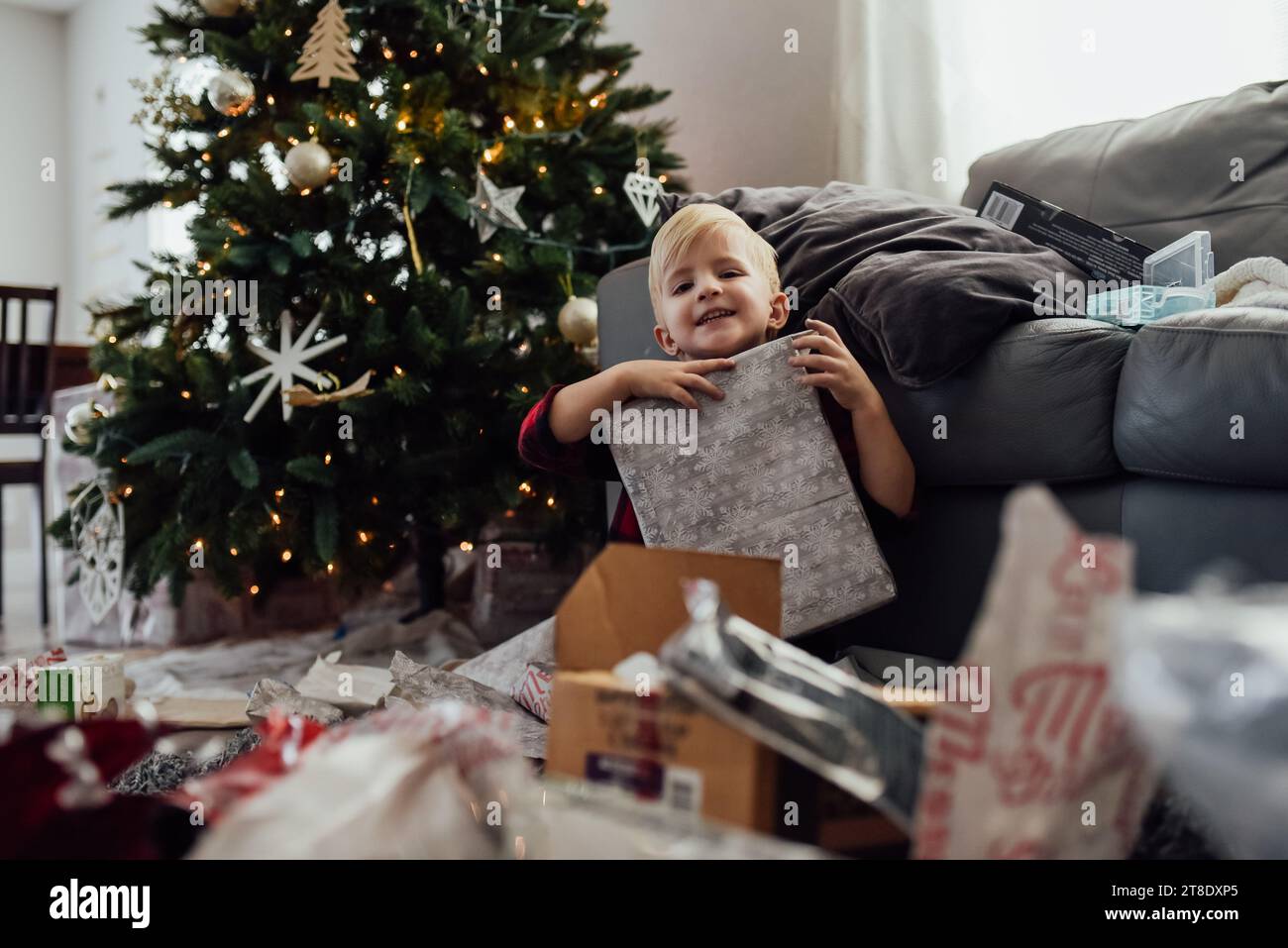 Small boy holds up present and smiles in front of christmas tree Stock Photo