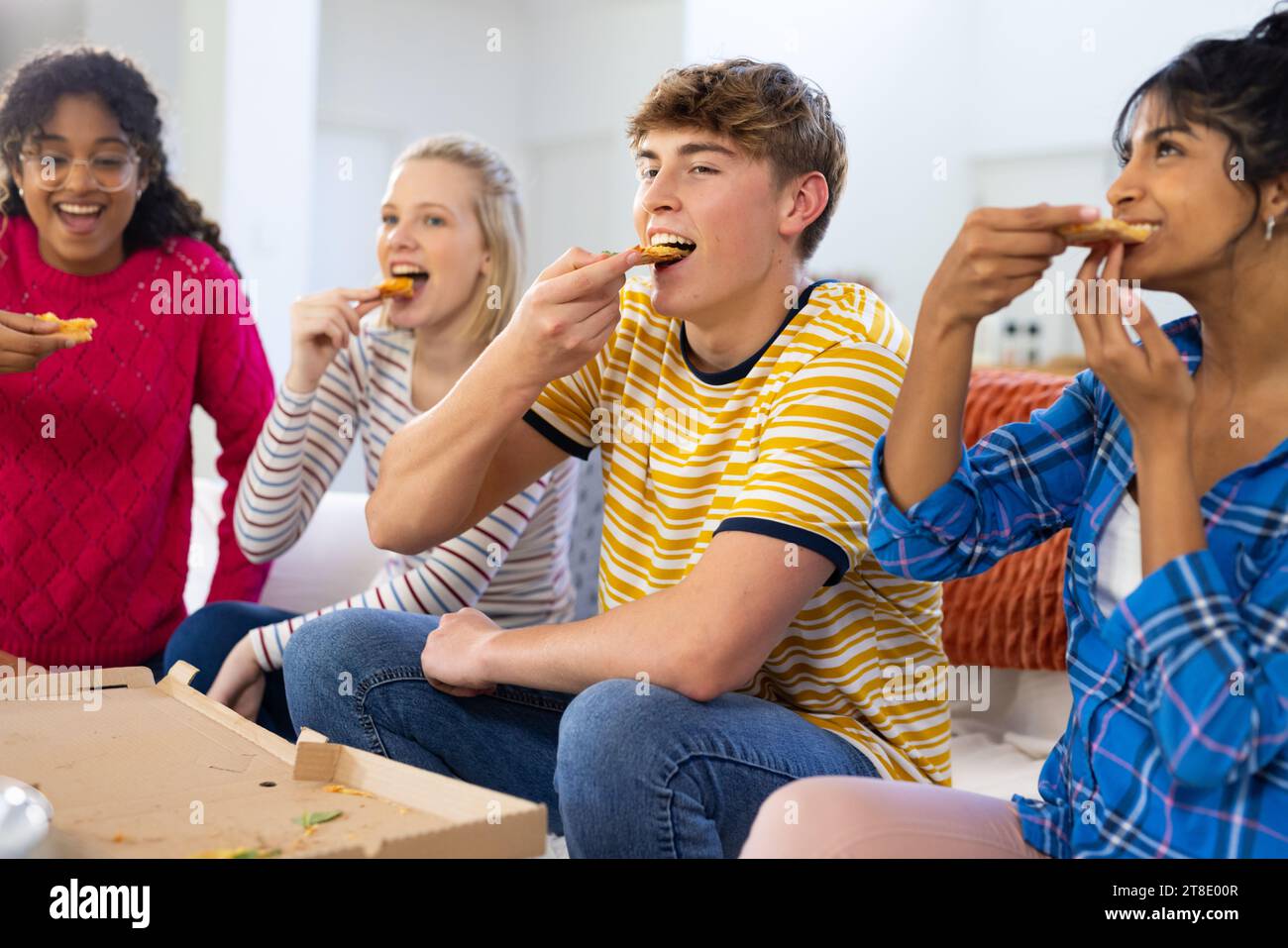 Happy diverse group of teenage friends sitting on couch and eating pizza Stock Photo