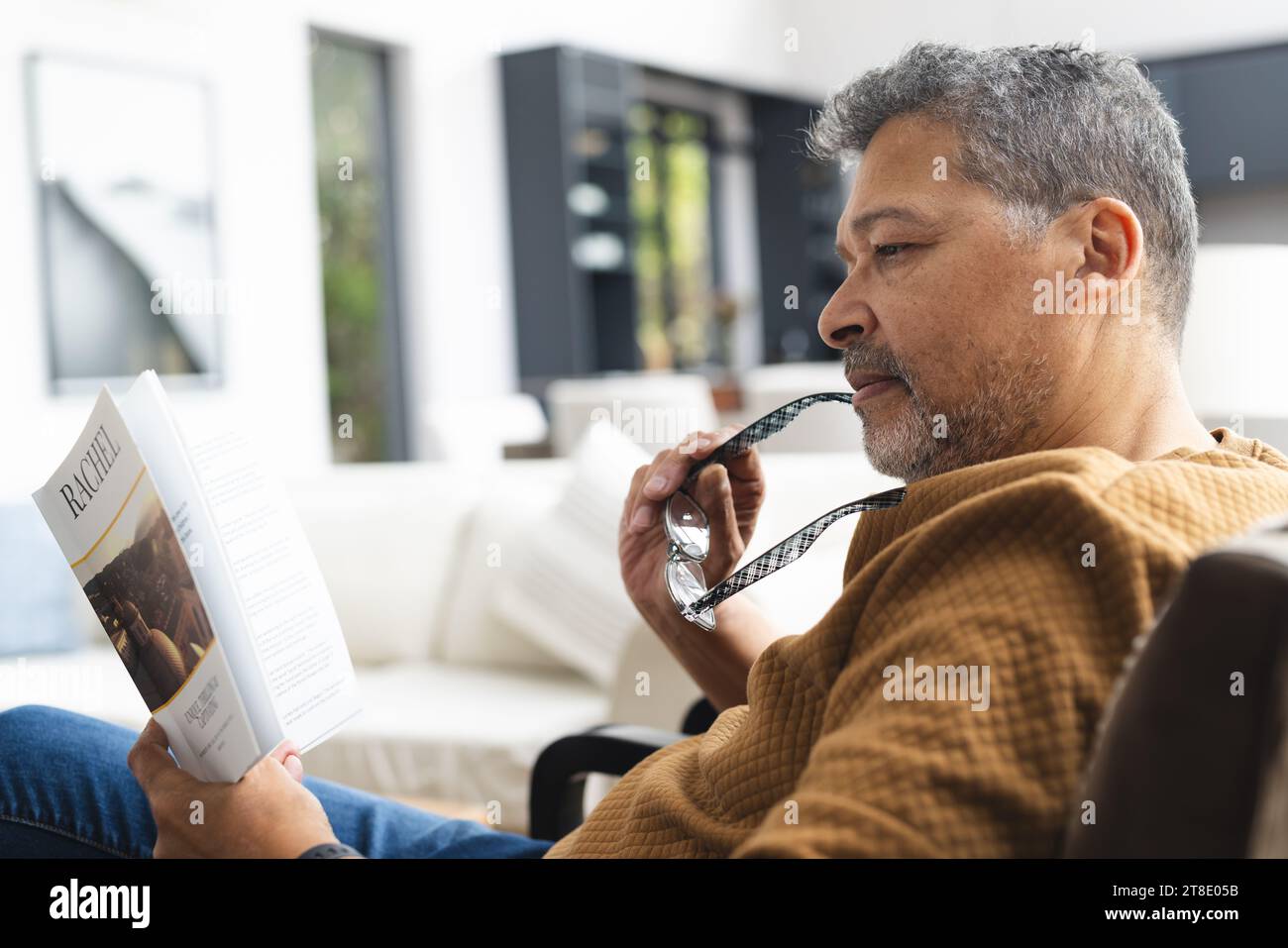 Happy senior biracial man sitting in armchair and reading book at home Stock Photo