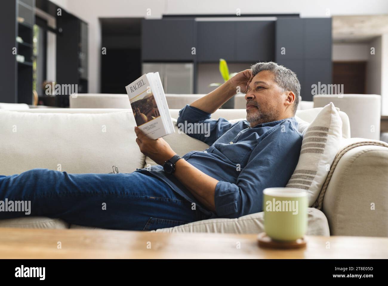 Happy senior biracial man lying on couch with cup of coffee and reading book at home Stock Photo