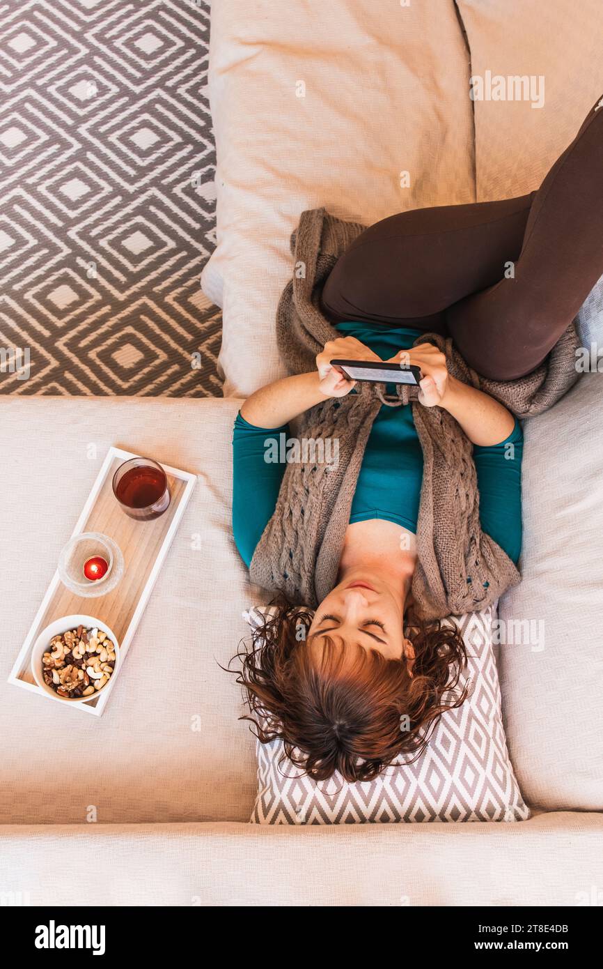 Vertical, woman, mid young, Caucasian, short haired, attractive, in casual clothes reading an ebook, lying on the sofa face up, in a Nordic style. Stock Photo