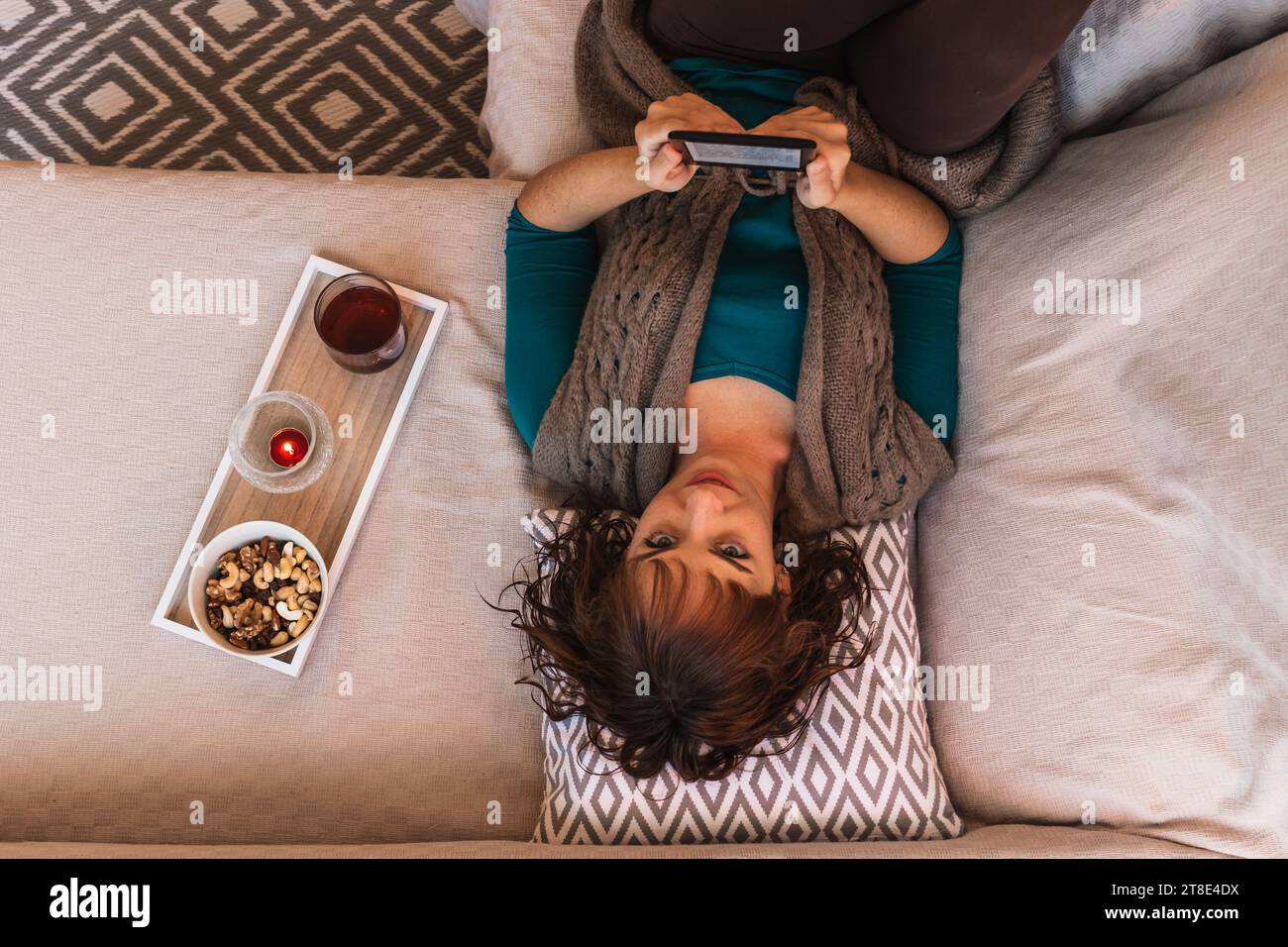 Horizontal, woman, mid young, Caucasian, short haired, attractive, in casual clothes reading an ebook, lying on the sofa face up, in a Nordic style. Stock Photo