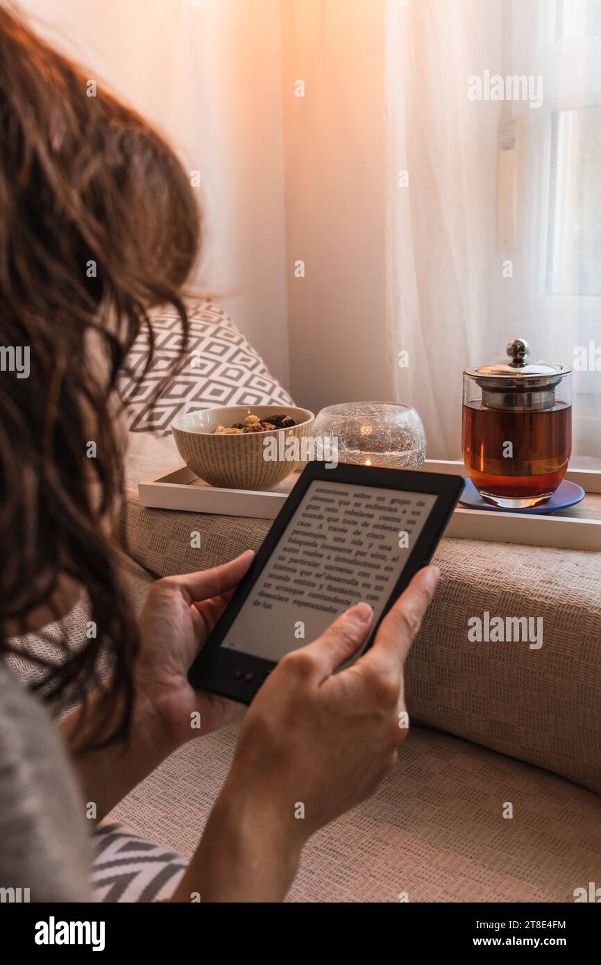 Vertical, mid young, Caucasian woman, with short curly hair, reading an ebook, accompanied by an infusion with nuts, lying on the sofa. Stock Photo