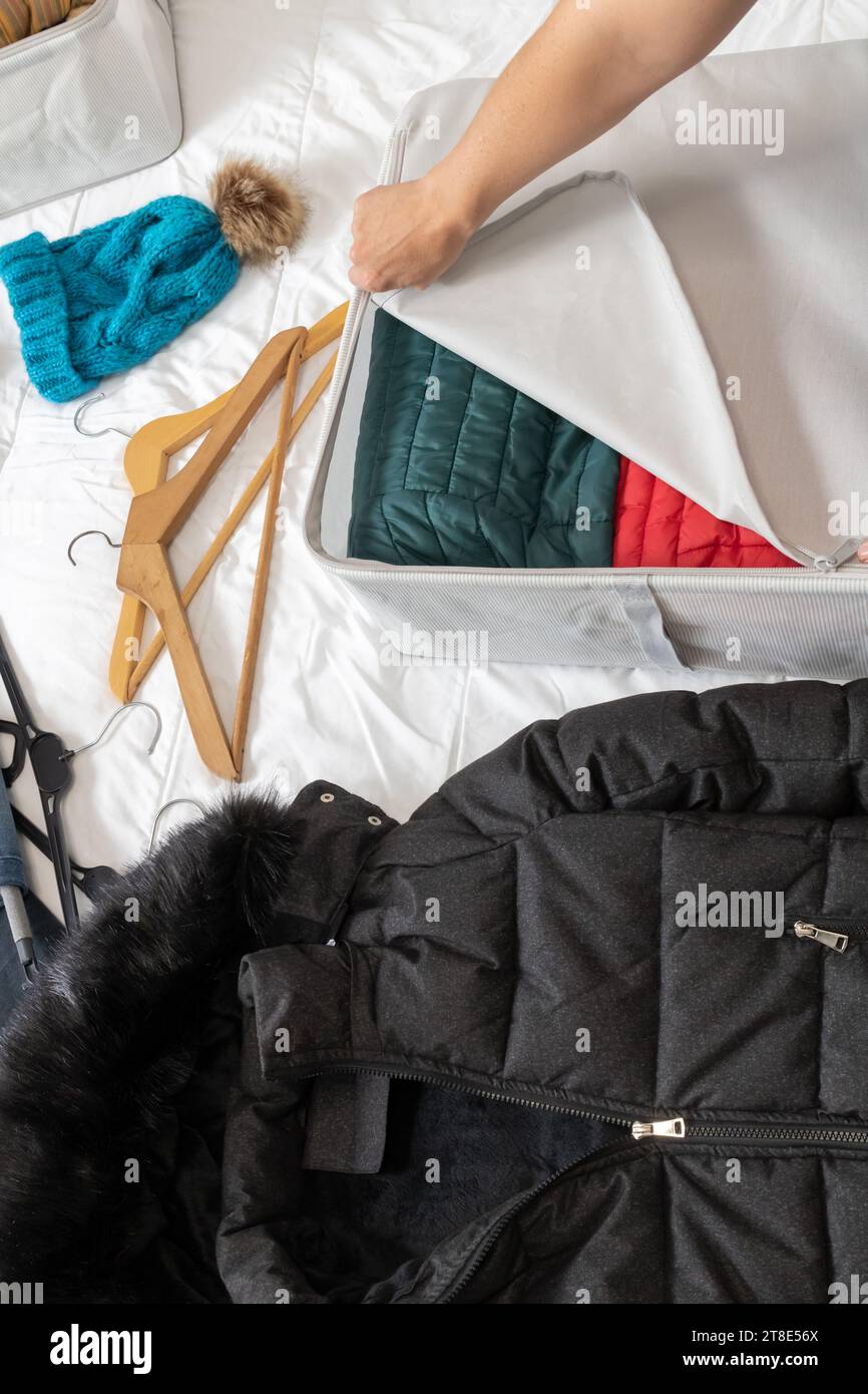 Vertical photo, woman's hand closing the zipper of a cloth box, with coats inside, on a bed, collecting winter clothes at the change of season. Stock Photo