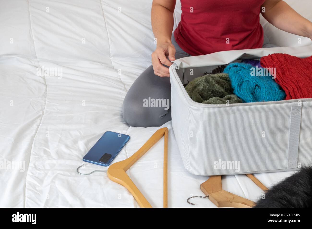 Horizontal photo, mid adult woman, Caucasian, with smartphone, sitting on top of bed, picking up winter clothes due to change of season. Stock Photo