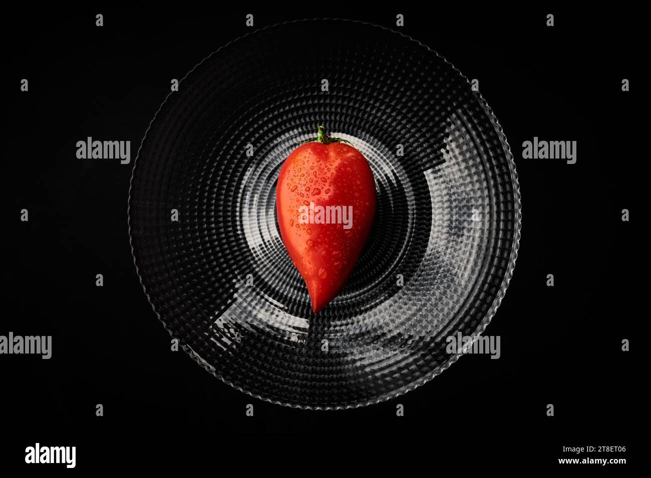Top view of a wet single tomato on a round, transparent plate with a black background, resembling a heart. Stock Photo