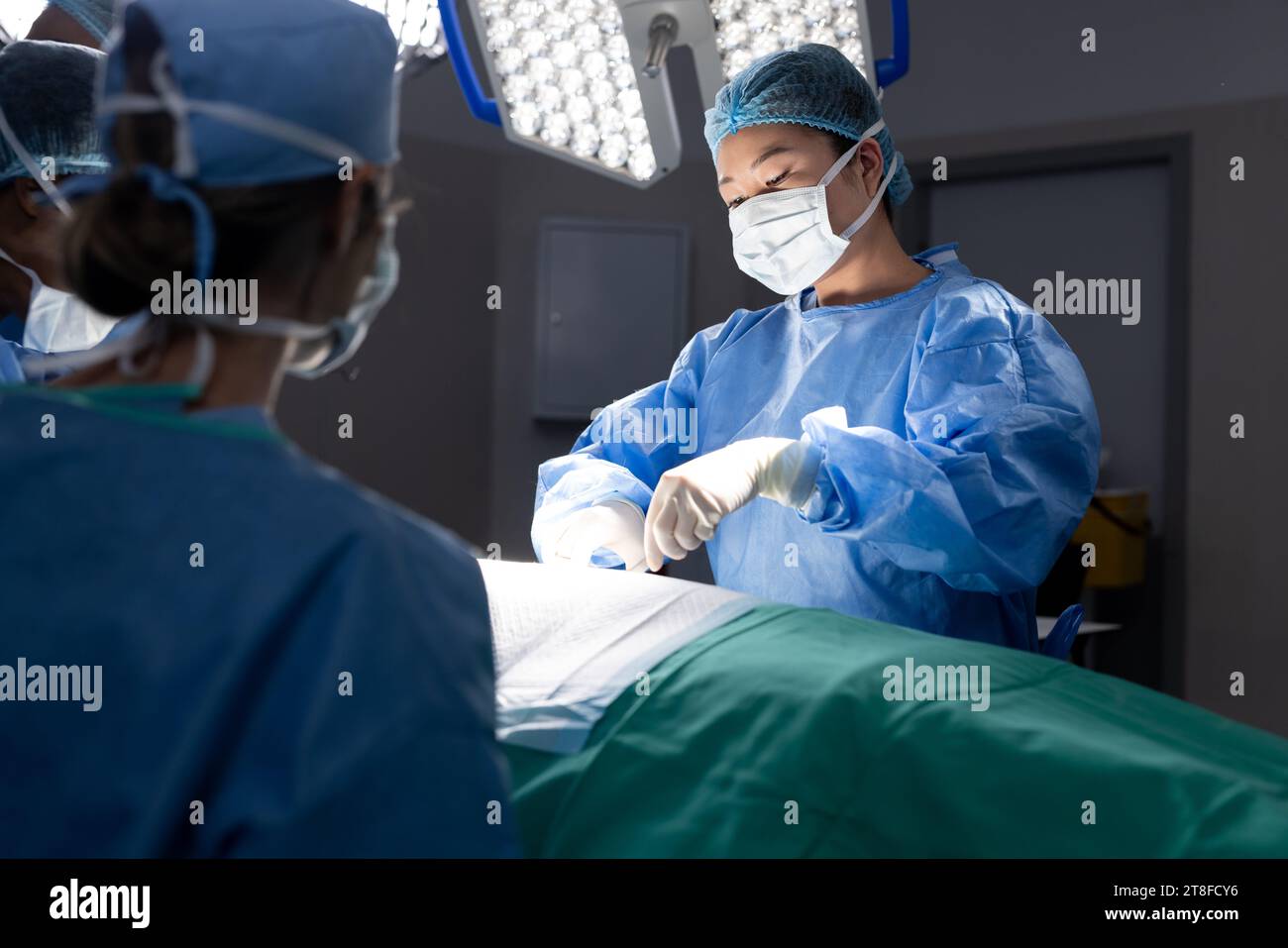 Diverse female doctor with face mask doing surgery in group of doctors in hospital operating room Stock Photo