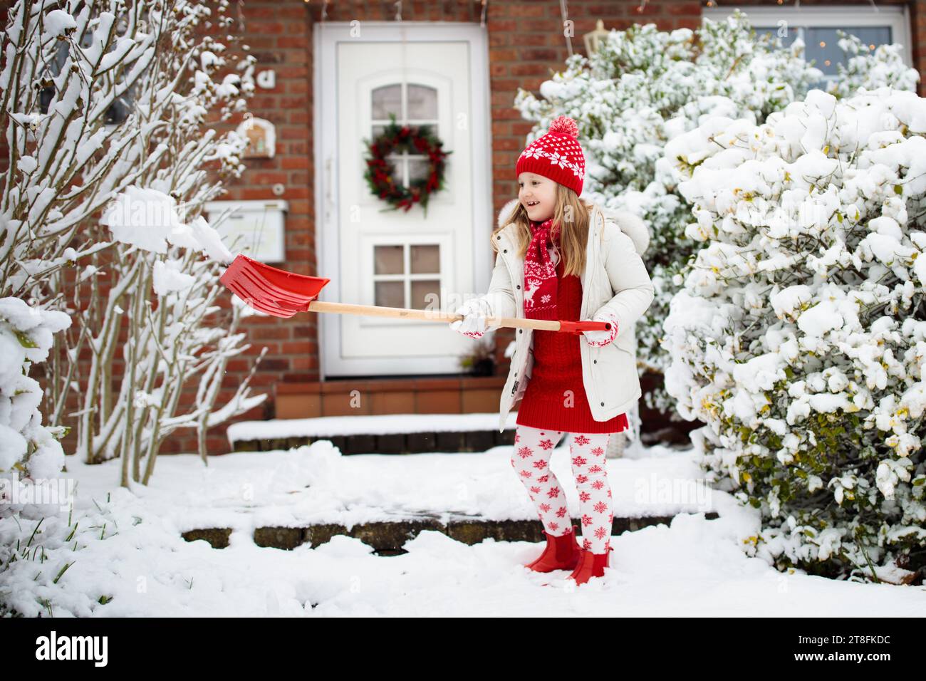Child shoveling snow. Little girl with spade clearing driveway after winter snowstorm. Kids clear path to house door after Christmas blizzard. Stock Photo