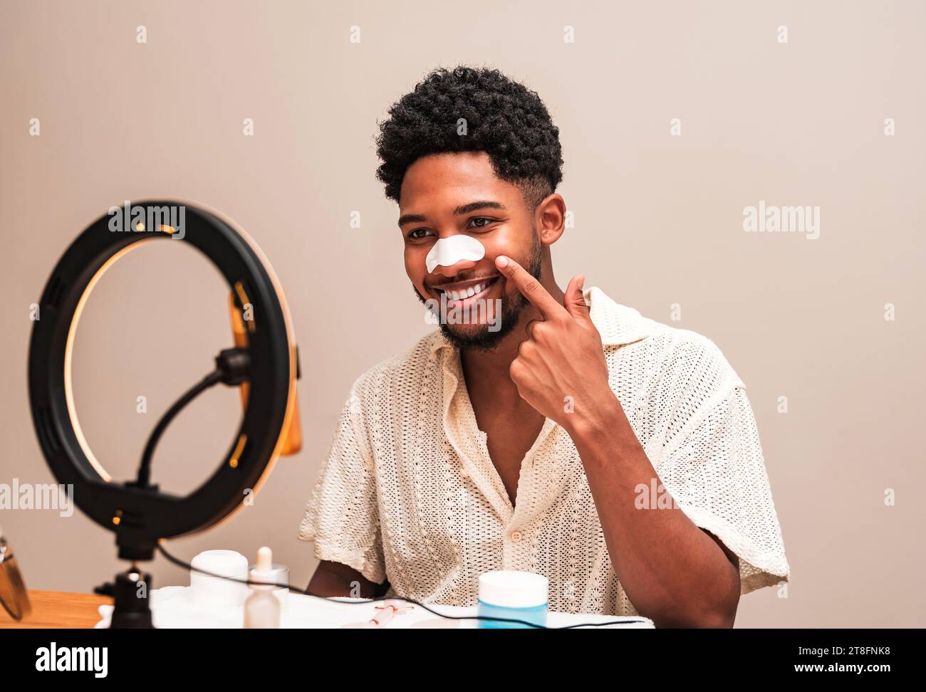 A cheerful young latin man applies a white nose pore strip while sitting at a table with skincare products and a ring light illuminating his face. Stock Photo