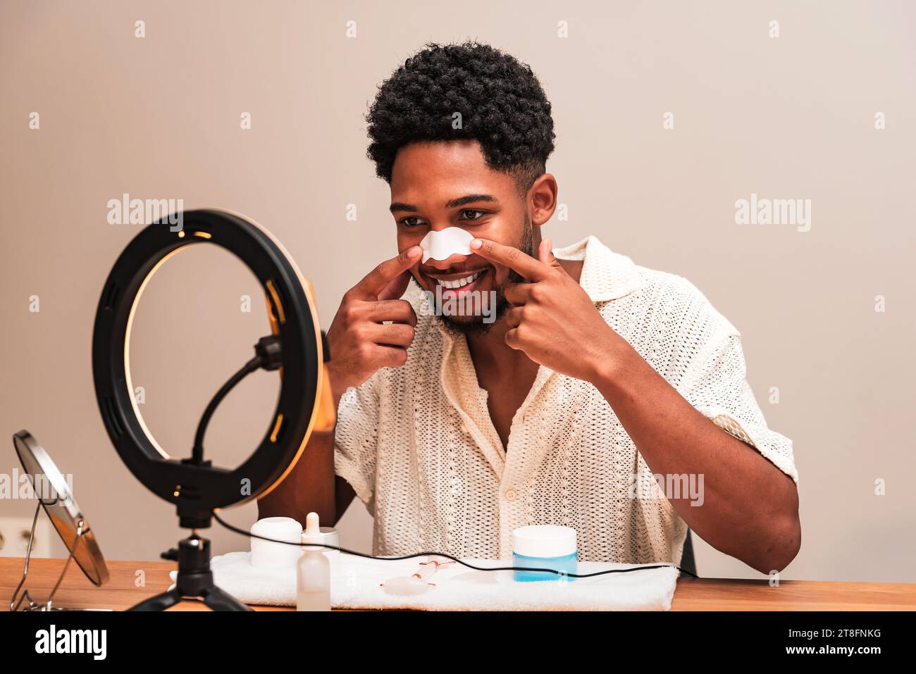 A cheerful young latin man applies a nose strip for skincare in front of a ring light, promoting self-care. Stock Photo