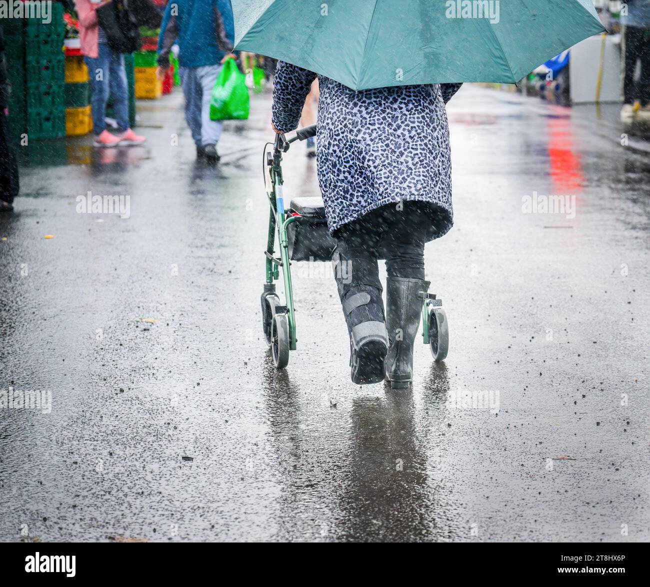 Woman wearing moon boot, pushing a mobility walker in the rain. Unrecognizable people shopping in the vegetable market. Stock Photo