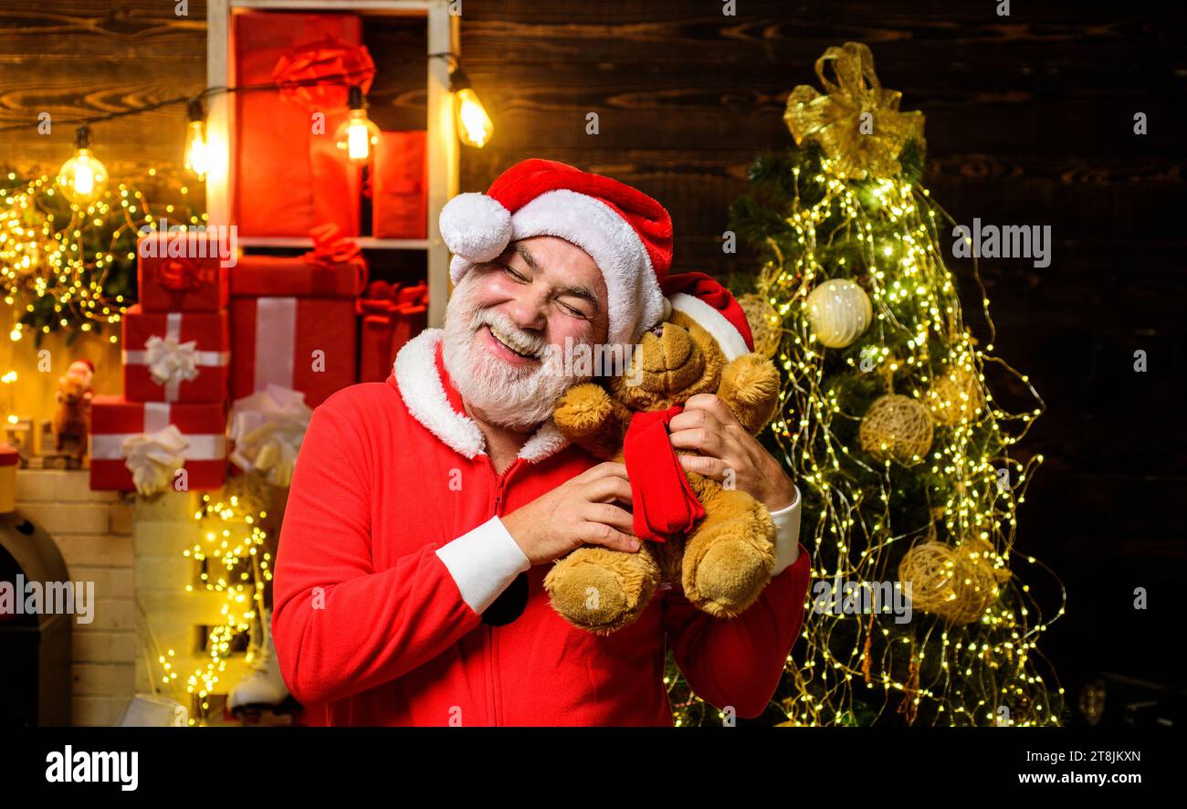 Happy Santa Claus hugs teddy bear. Cheerful bearded man in Santa Claus costume with plush toy teddy bear. Santa man with teddy bear in room decorated Stock Photo