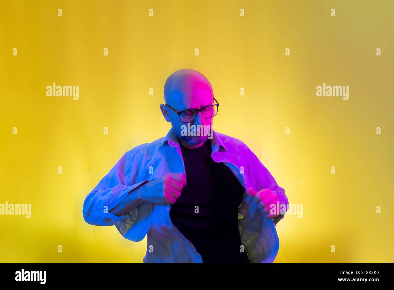 Bearded, bald man with glasses opening his shirt. Isolated on yellow background. Stock Photo