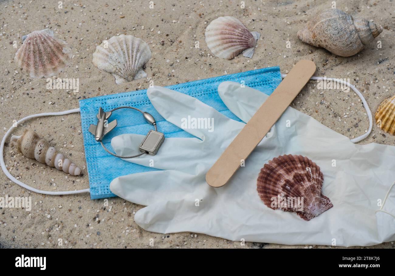 face mask, latex glove, spatula and luggage tag with plane, with shells and snailsshells on the beach, summer holidays under Corona conditions Stock Photo