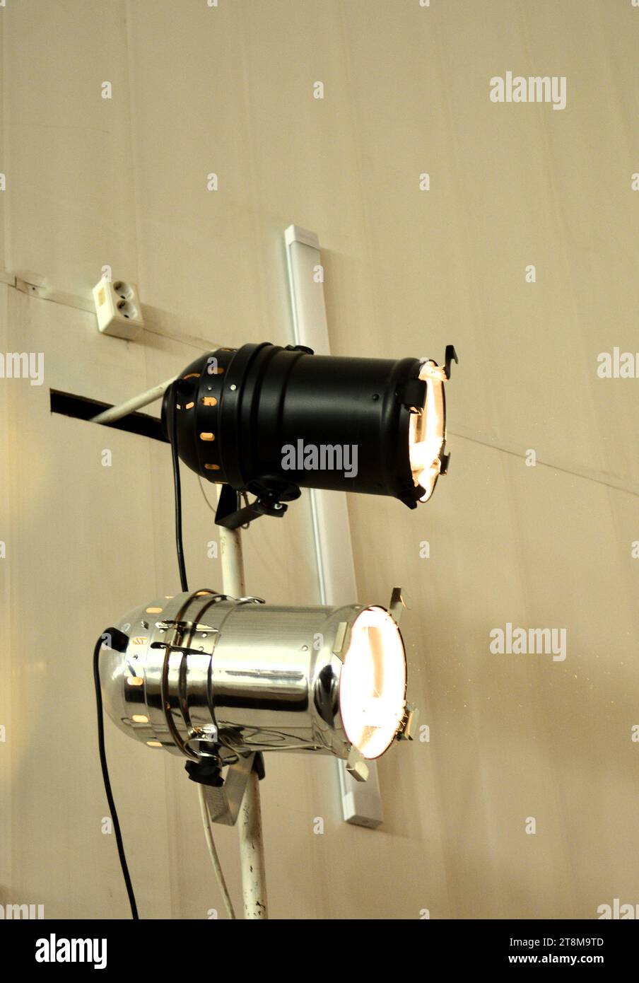 Two turned on theater spotlights on a white wall with wires and an electrical outlet. Close-up. Stock Photo