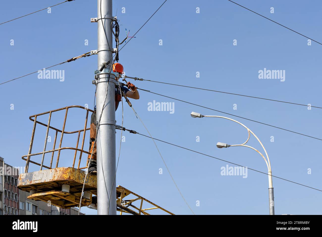 A worker tensions and secures a cable on a lamp post while standing in a lifting construction basket against a blue sky. Stock Photo