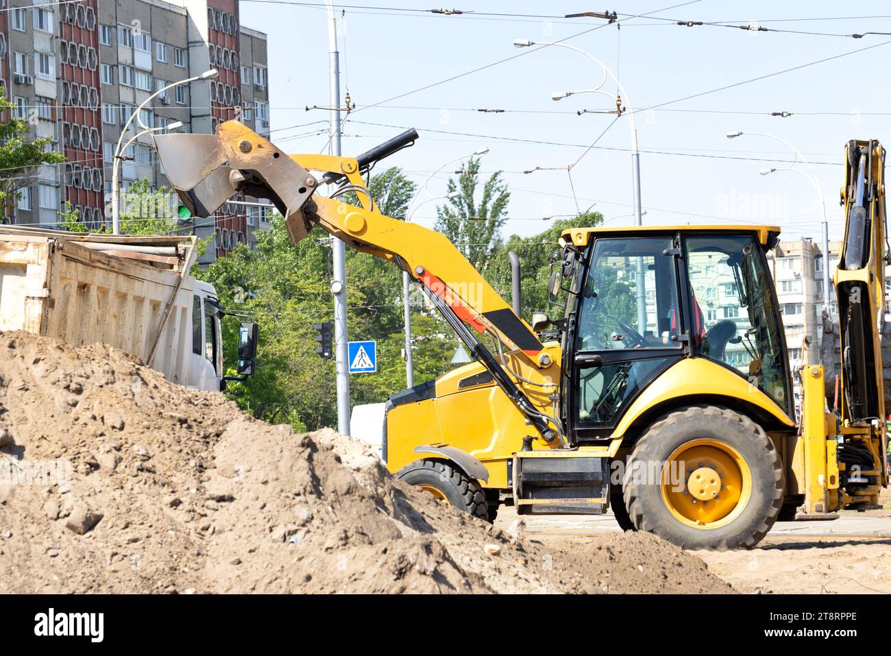 The sliding bucket of a road excavator works to load sand into the back of a truck against the backdrop of a city street on a summer day. Stock Photo
