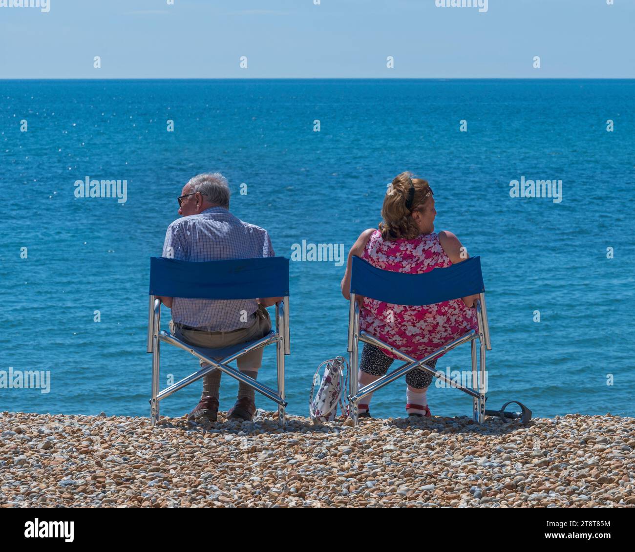A couple sat on the stone beach in their fold up chairs, facing away from each other on the beach at Lyme Regis, Dorset, England, UK Stock Photo