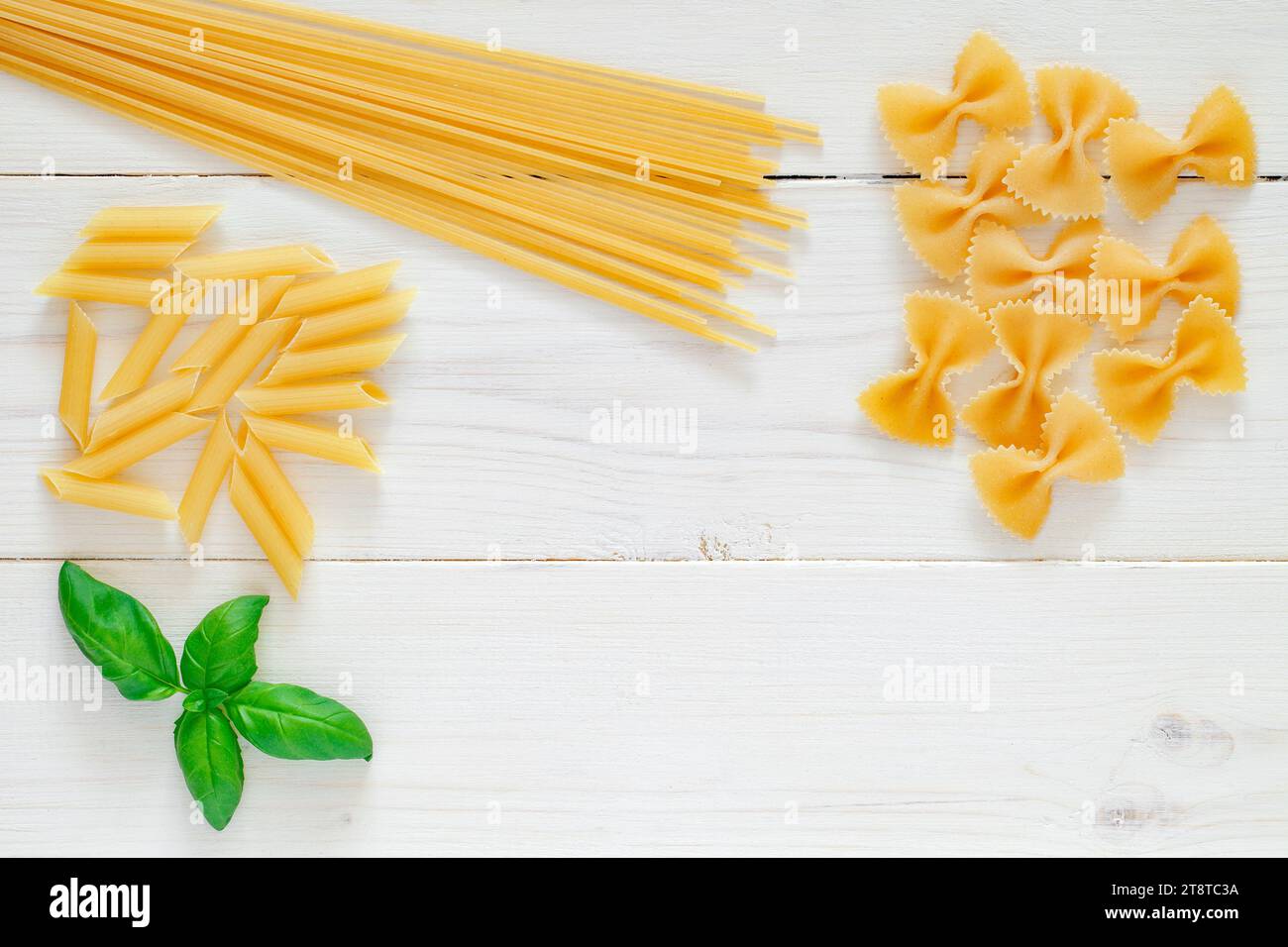 Maccheroni raw, spaghetti, farfalle, pipe, rigatoni, penne, rigate, leaf of basil, on white wooden board background, top view, space to copy text. Stock Photo