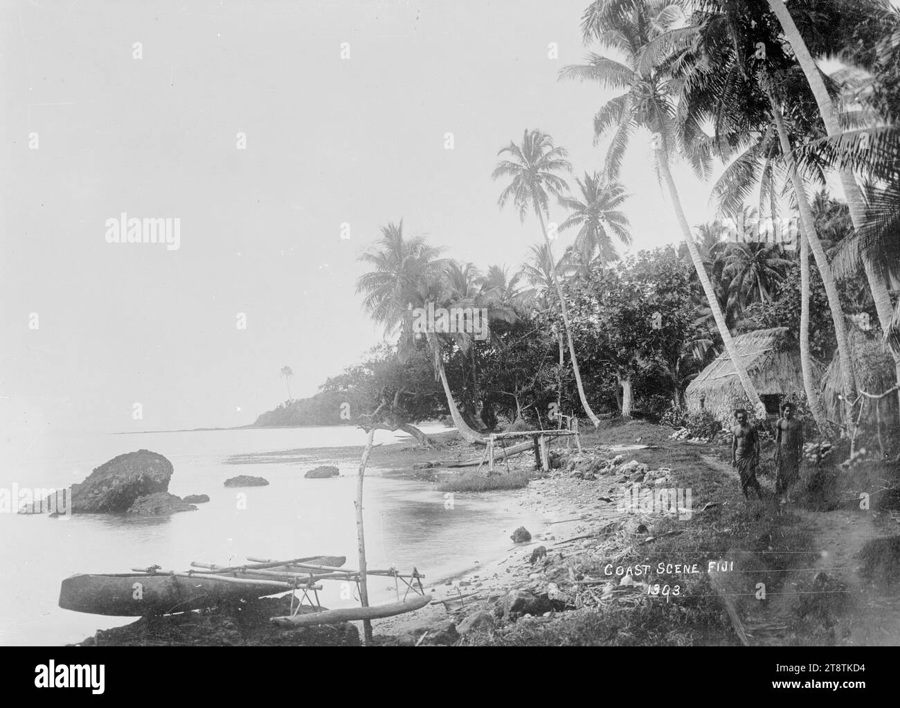 Coastal scene, Ovalau, Fiji, View of a beach fringed with palm trees. An out-rigger canoe is drawn up at the water's edge in the foreground. There is a hut near the foreshore and two Fijian men standing near by looking at the photographer. Taken in early 1900s Stock Photo