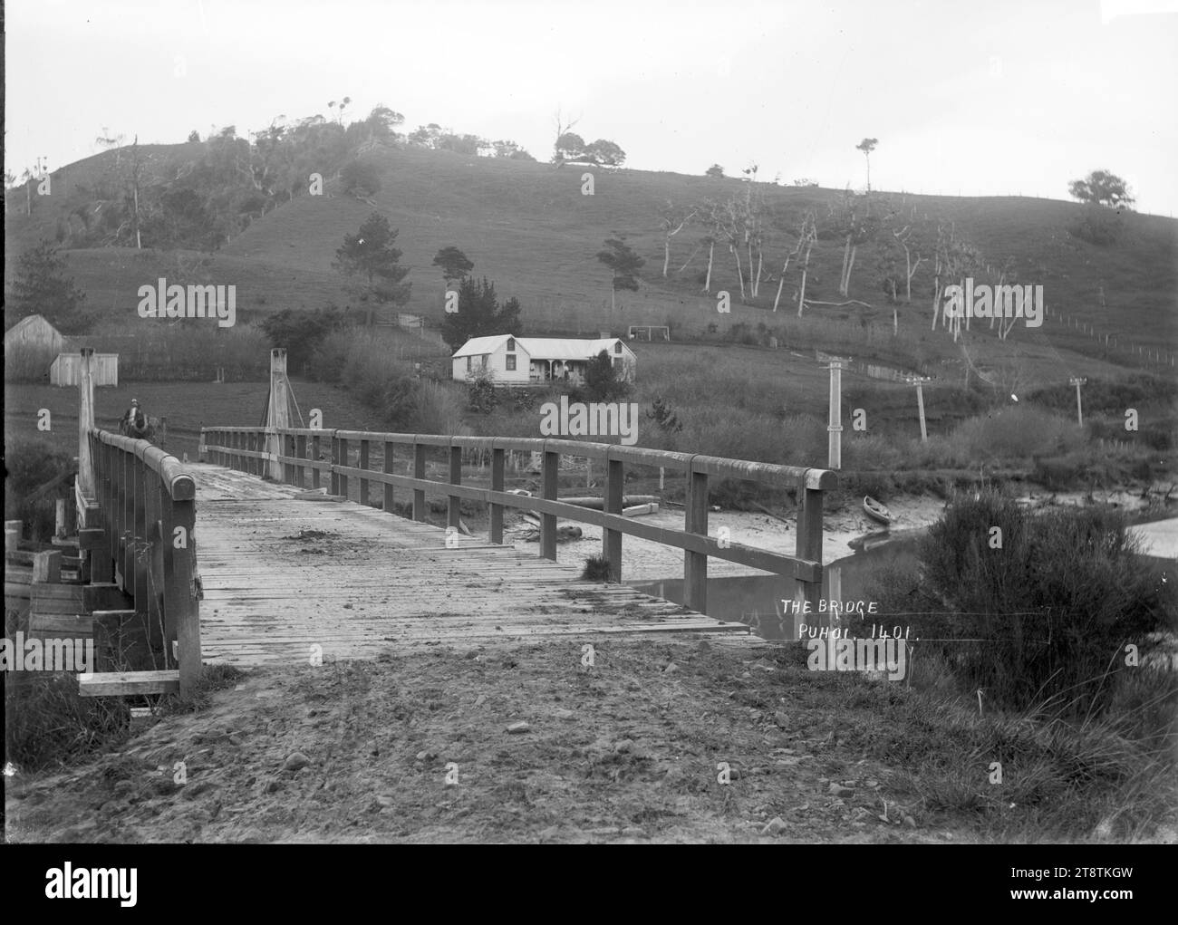 Bridge across the Puhoi River, Puhoi, View of the river and wooden bridge. There is a house and telegraph posts across the river on the opposite bank. A man on a horse is at the far end of the bridge. Photograph taken 1900-1930 Stock Photo