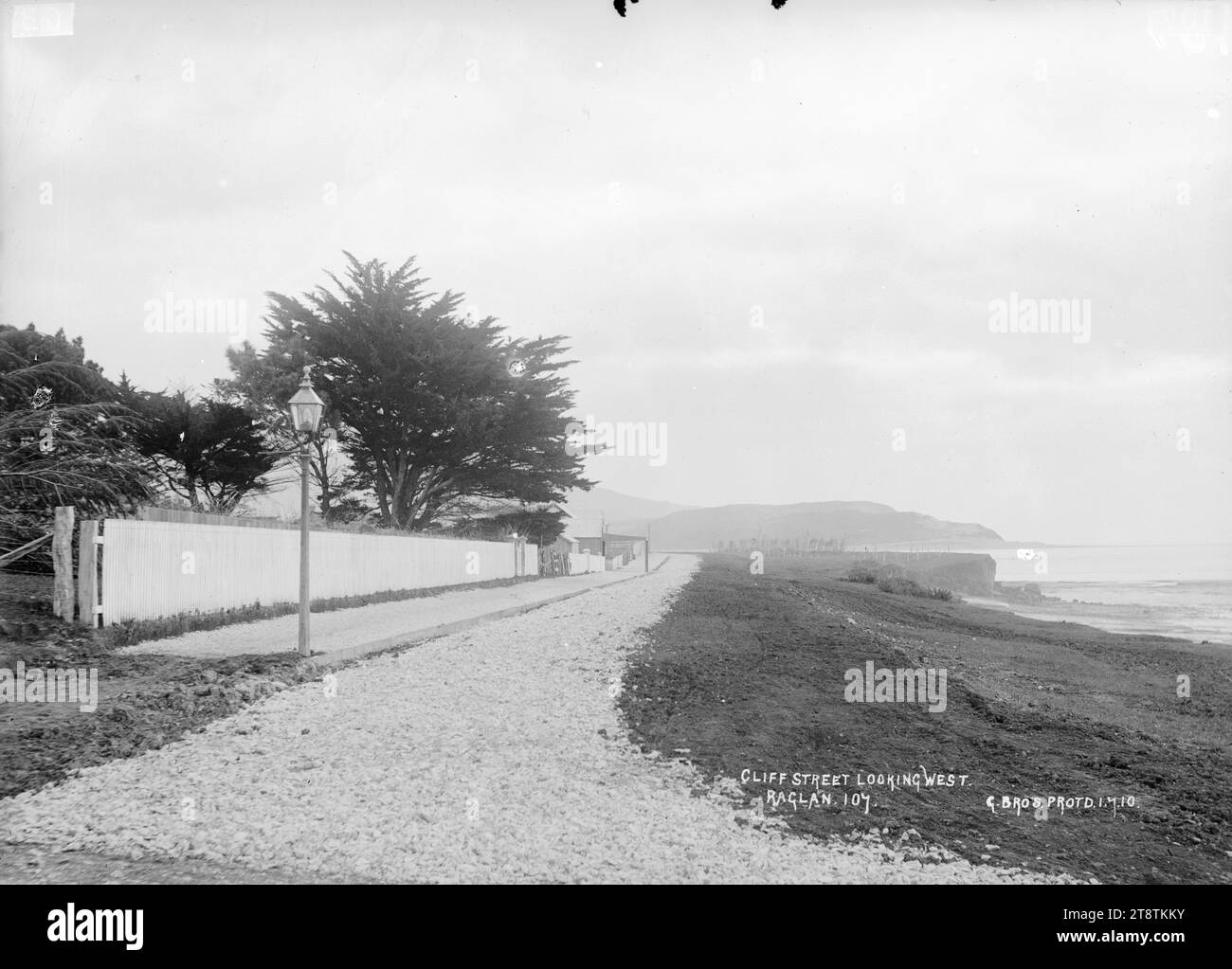 Cliff Street, Raglan, New Zealand, 1910, Cliff Street, Raglan, New Zealand, looking West. The street runs between the coast and a white picket fence. A street lamp is at the edge of a footpath near the fence. Photograph taken by the Gilmour Brothers Stock Photo