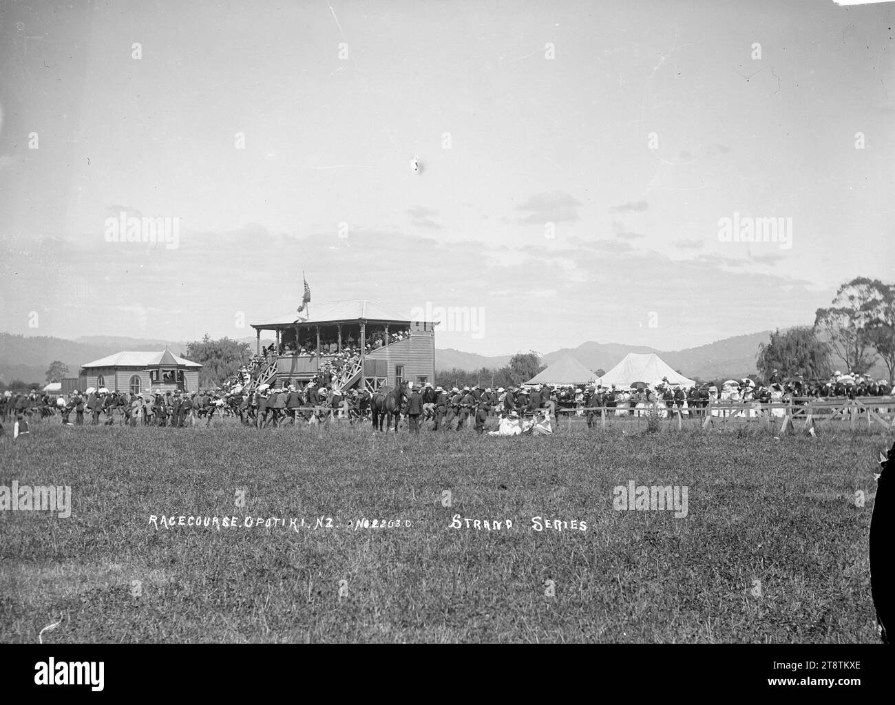 Grandstand and racecourse, Opotiki, View of the grandstand, buildings, tents on race day at the Opotiki race course. Spectators are viewing the racing from the grandstand and lining the fences close to the stand. in early 1900s Stock Photo
