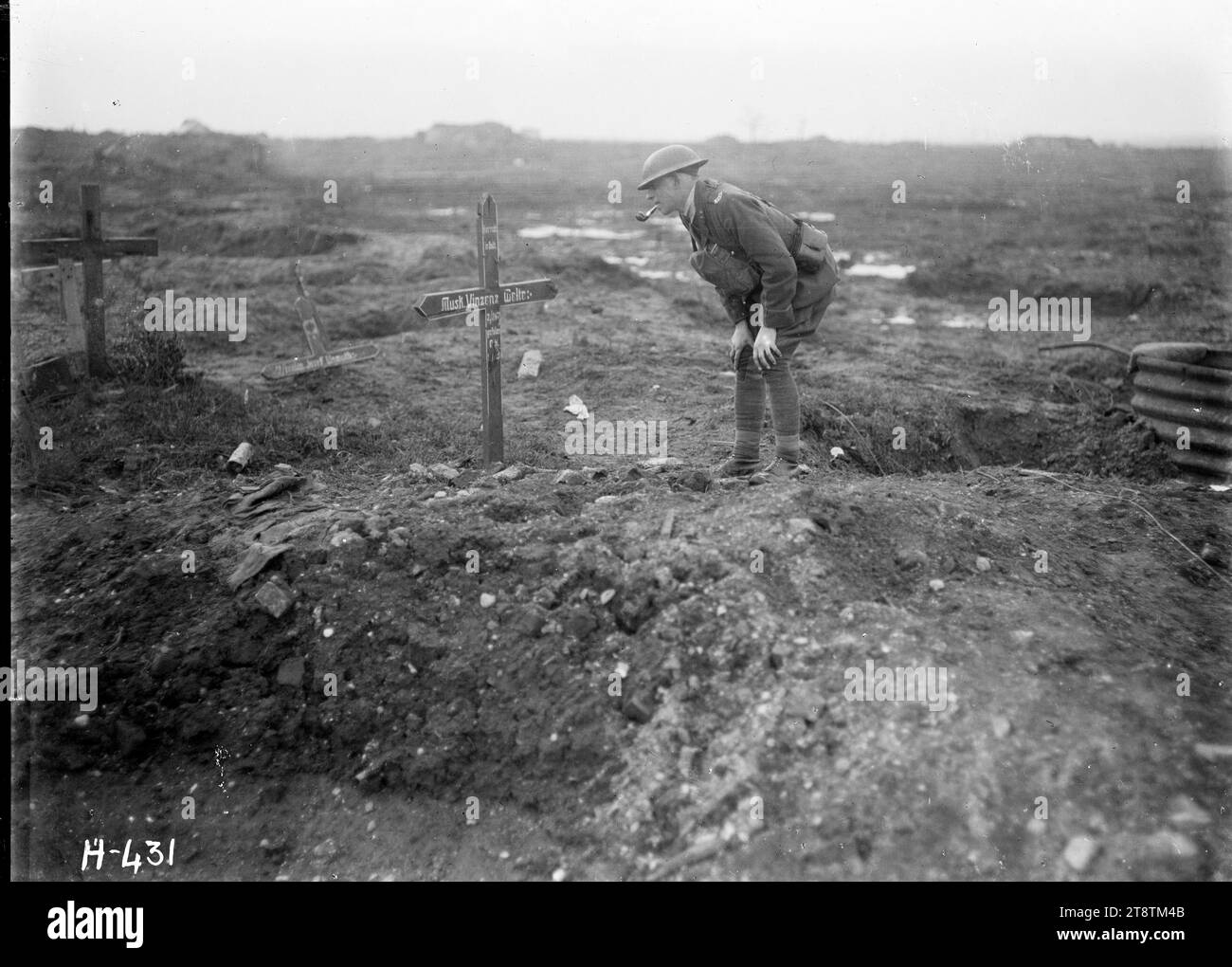 A New Zealand officer reads a cross in a former German field cemetery, World War I, A New Zealand Artillery officer bends over to read the wooden cross marking a grave in a former German field cemetery that is now a target of German shelling in World War I. Water-filled shell holes can be seen in the background. There is also a broken wooden cross on the ground. Photograph taken at Westhoek Ridge 4 February 1918 Stock Photo