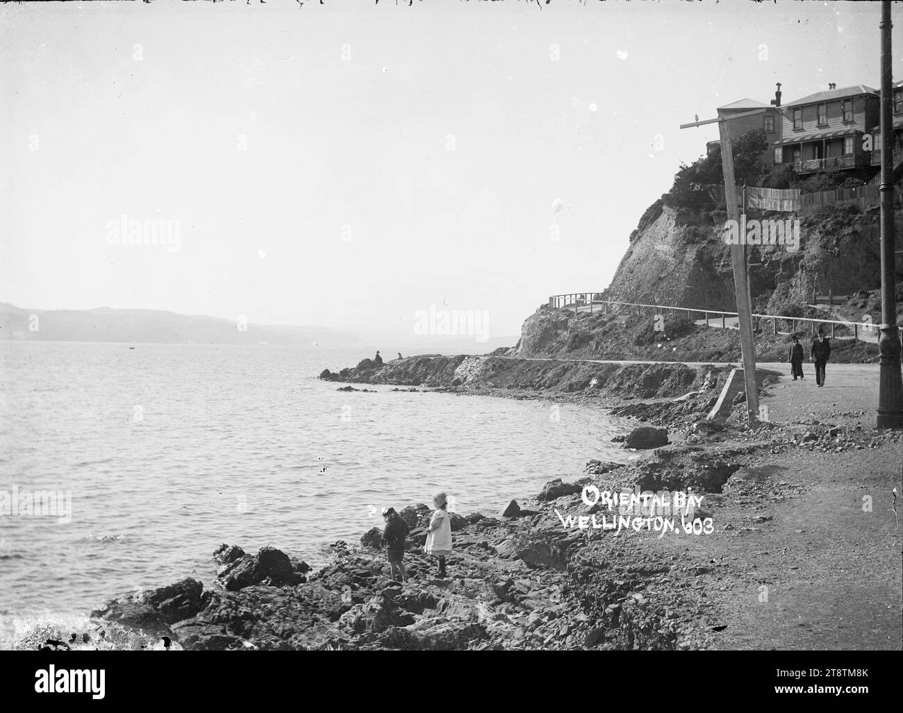 Oriental Bay, Wellington, New Zealand, View taken from Oriental Parade with Carlton Gore Road on the right and Lambton Harbour on the left. Two children are standing on the rocky foreshore in the immediate foreground. Two men are on Oriental Parade on the right. Houses overlooking Oriental Bay (on Carlton Gore Road) are visible in the top right. Photograph taken between 1910 and 1930 Stock Photo