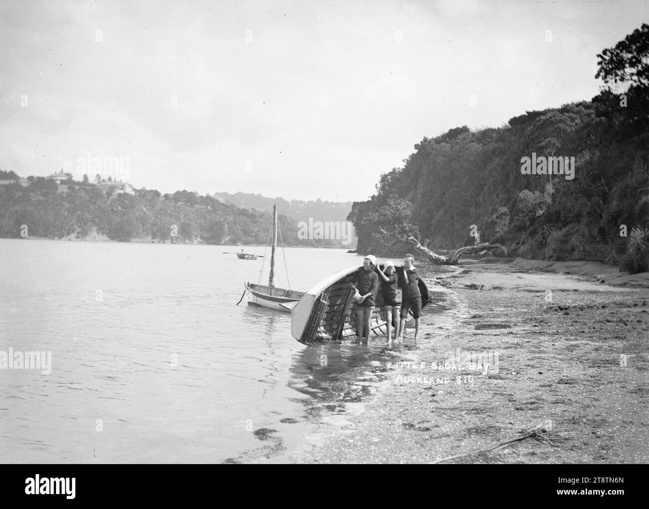 Boys with a dinghy at Little Shoal Bay, Auckland, New Zealand, Three boys holding a dinghy on its side at the water's edge in Little Shoal Bay, Northcote. Anchored nearby is a small sailing boat. Further off-shore is a man rowing a dinghy. in early 1900s Stock Photo