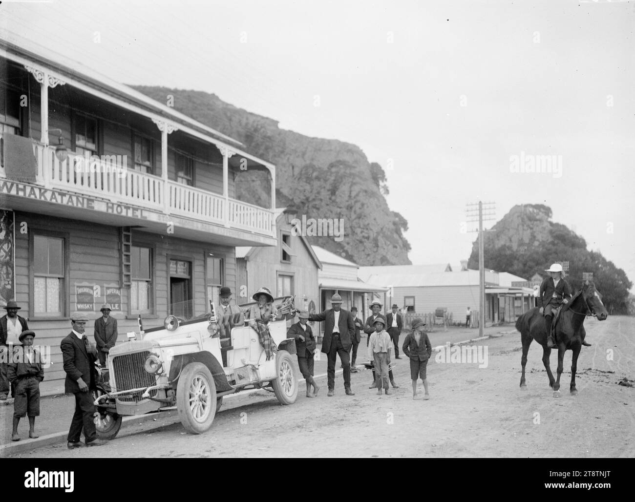 Whakatane township, Whakatane Hotel with rocky ciffs behind, photographed circa 1910s. Looks along a street with the Whakatane Hotel on the left. A touring car with a man and a woman as passengers in the back seat, with a group of people standing around, and a young man on horseback, watching from the middle of the road Stock Photo