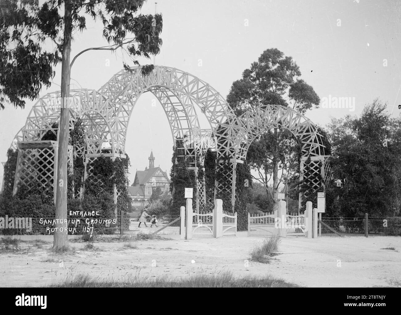 Princes Gate at the entrance to the grounds of the Government Sanatorium and Baths at Rotorua, View of the triple archway and flagpole (known as Princes Gate) at the entrance leading from Hinemaru Street to Queens Drive, and to the gardens of the Government Sanatorium and Baths. There are wooden gates at the entranceway and a wire fence. There are noticeboards on top of the fenceposts and another close by. The Tudor Towers bathhouse (built 1906-1907) can be seen in the distance. A girl pushing a pram and a young child are in the gardens just inside the entranceway. Stock Photo