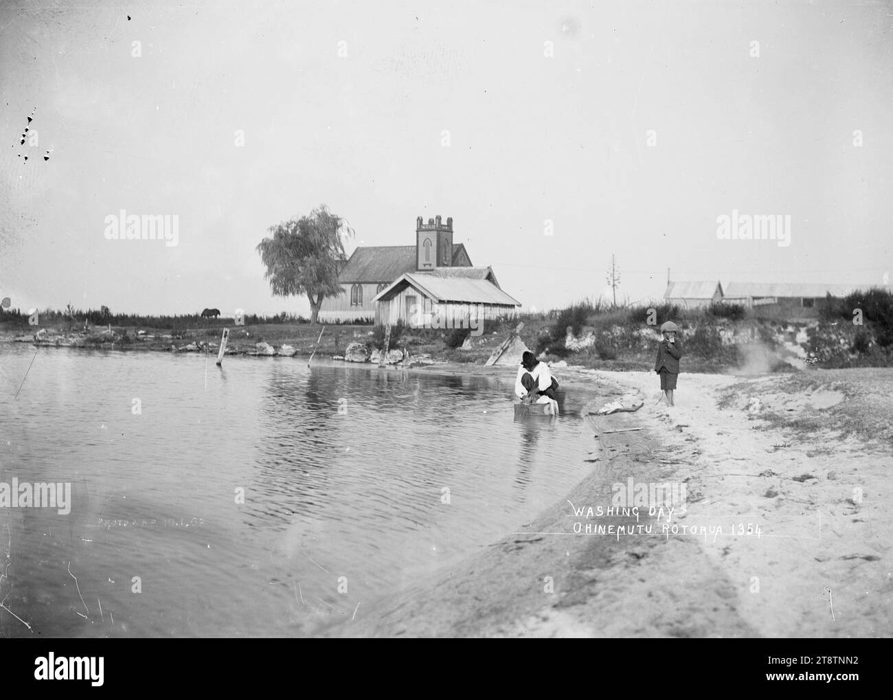 View of St Faith's Church, Ohinemutu, View of St Faith's Church (old church), at Ohinemutu, taken from the lakeside looking south. In the foreground a Maori woman is washing clothes at the lake edge while a young boy stands nearby. Photograph taken in early 1900s Stock Photo