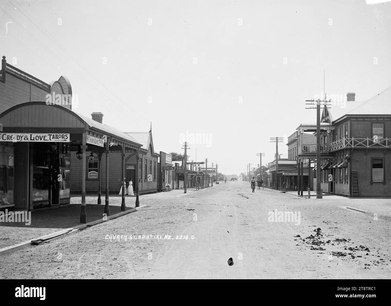 Church Street, Opotiki, View looking down Church Street (now known as Elliot Street) with the Royal Hotel on the right; McCready & Love (tailors), Mechanics Institute; dentist, saddler, and other businesses on the left. The Masonic Hotel can be seen in the far distance on the left. Two women are standing at the entrance to the Mechanics Institute; a pram is on the footpath outside the dentist; a cyclist is heading towards the photographer and horseriders can be seen in the far distance. in early 1900s Stock Photo