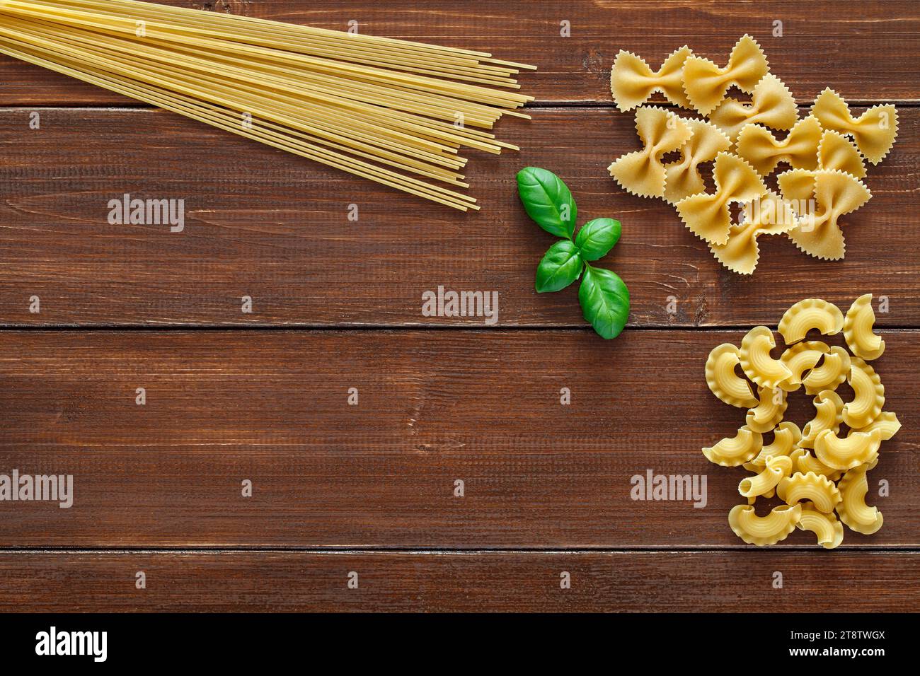 Spaghetti, maccheroni raw, farfalle, cresta di gallo, and green leaves of basil on wooden brown planks dark background, top view, space to copy text. Stock Photo